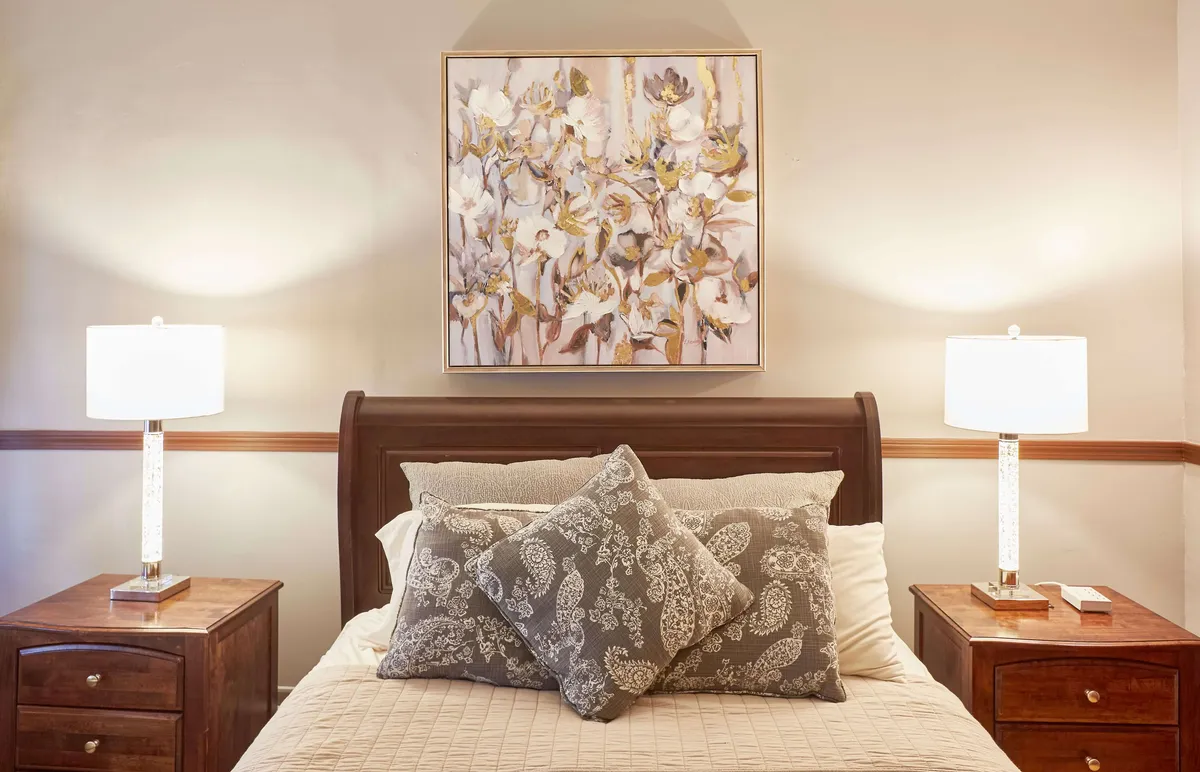This bedroom scene includes a plush bed adorned with decorative pillows, warm table lamps on nightstands, and artwork above the headboard. The lighting and soft textures create a tranquil retreat ideal for rest and reflection.