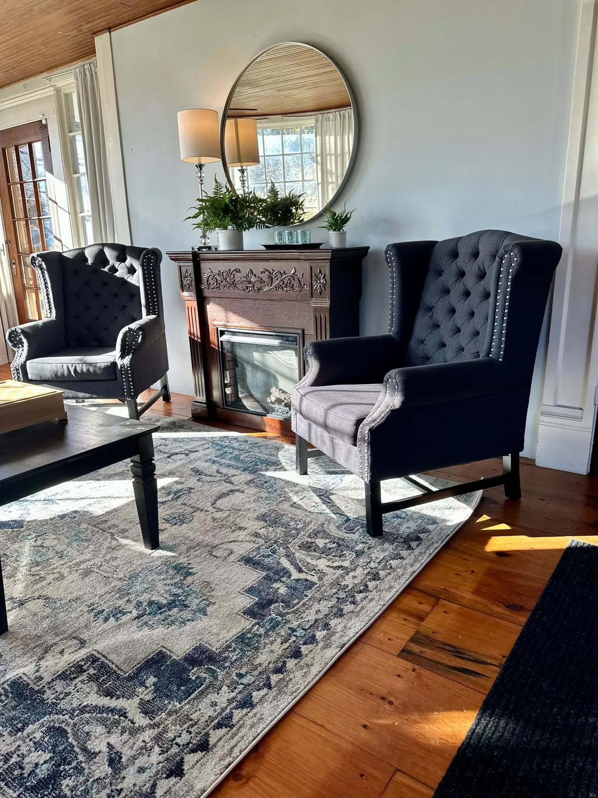 This interior shot captures two upholstered armchairs positioned near an ornate fireplace, a decorative area rug, a large round mirror, and lush potted plants. The mood is cozy and celebratory, perfect for sharing drinks or casual conversations with friends and family.
