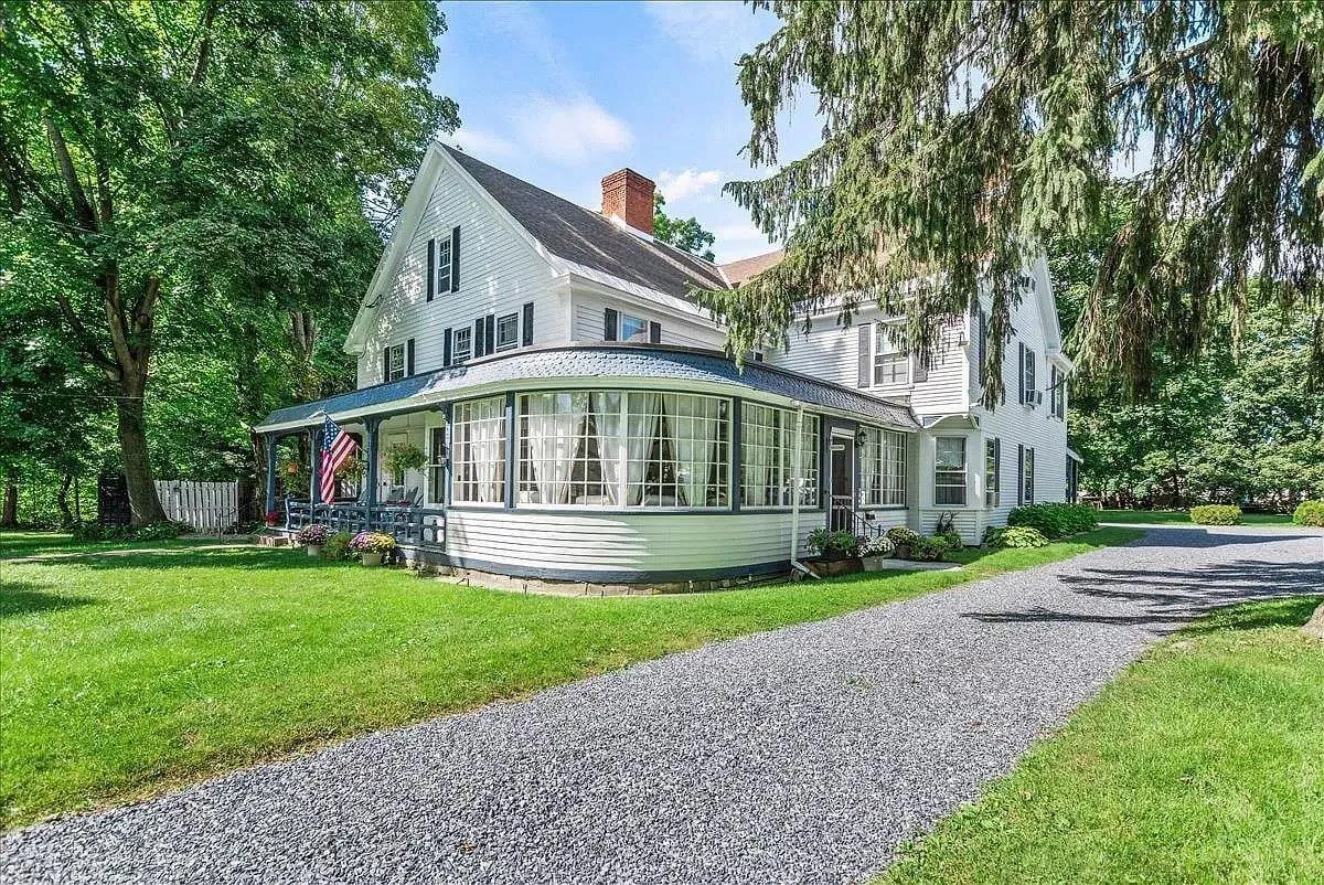The image showcases a stately historic home with a spacious wraparound porch, elegant bay windows, and a lush, well-kept lawn. The inviting porch is perfect for relaxing with morning coffee or enjoying a serene sunset, while the manicured garden offers a peaceful retreat just steps from the vibrant heart of downtown Bennington.