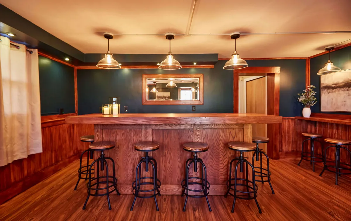 A stylish bar area featuring a wooden bar, metal stools, hanging light fixtures, a wall mirror, and a decorative plant. The setup is ideal for hosting social gatherings or casual brunches, reflecting a space where guests mingle and enjoy each other's company.