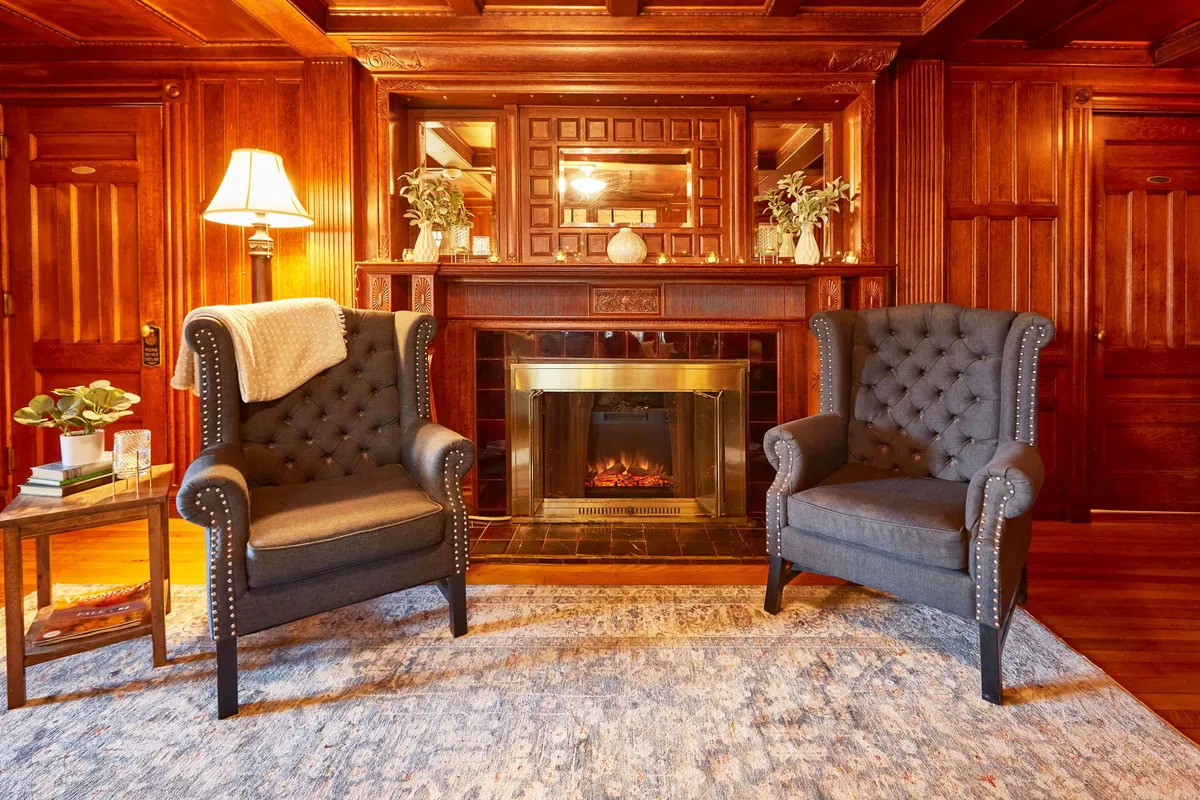 A pair of tufted chairs sit intimately beside a roaring fireplace, set against warm wood paneling. The room is styled for comfort, with soft textiles and ambient lighting, suggesting a perfect spot for quiet conversation or enjoying a glass of wine by the fire.
