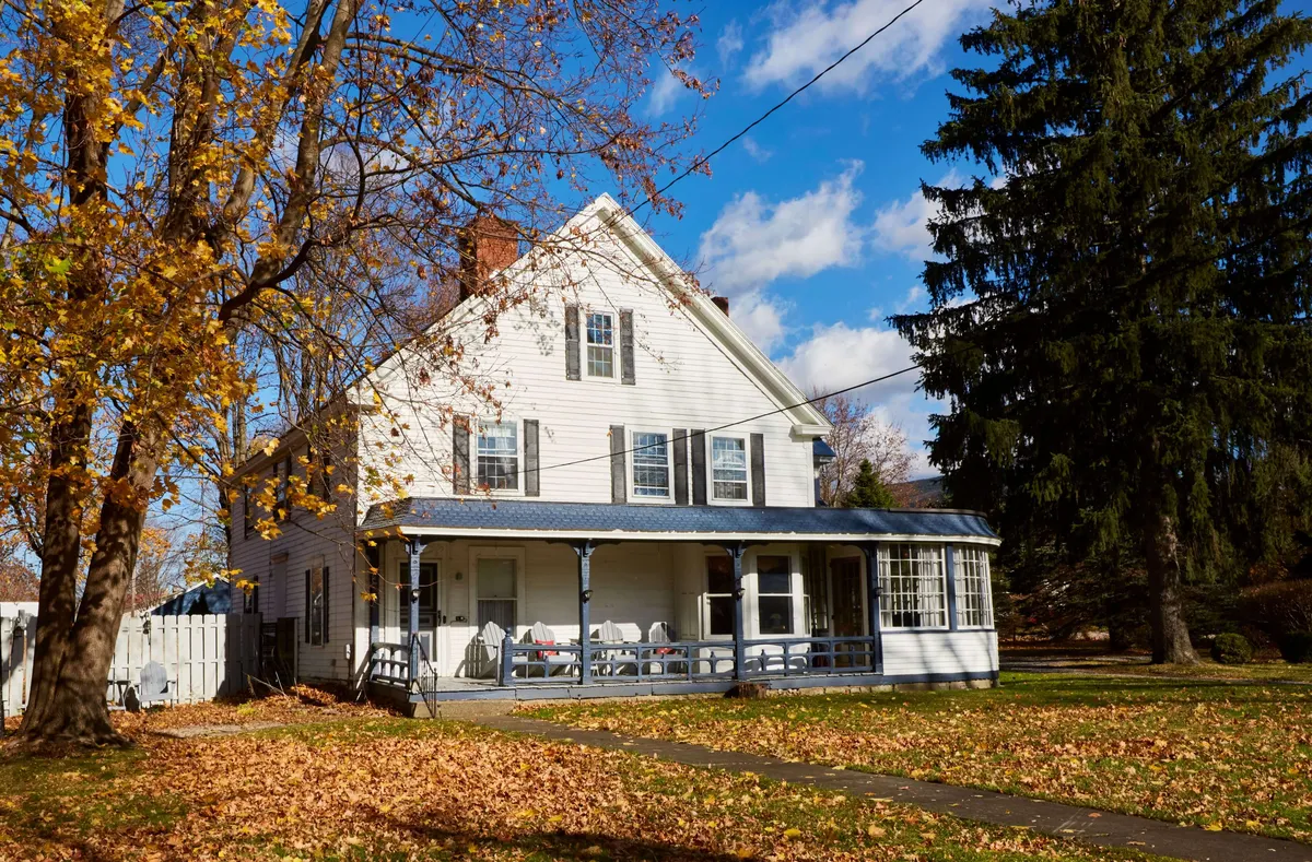 A welcoming exterior view of the house's front porch, framed by large windows and surrounded by vibrant autumn leaves. The porch offers a comfortable space for enjoying coffee or relaxing outdoors, with the home's classic New England charm on full display.