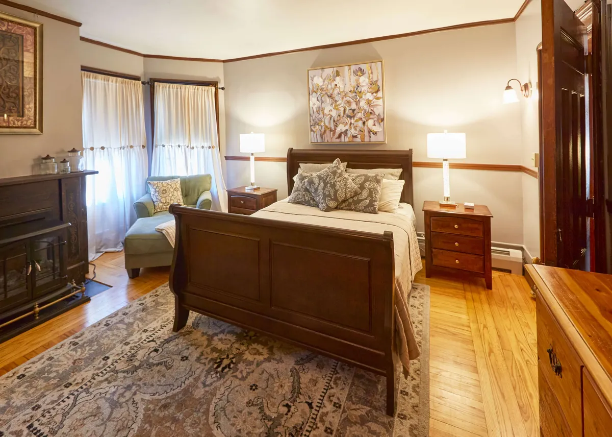 A warmly lit bedroom featuring a comfortable bed, plush armchair beside a glowing fireplace, and tasteful artwork. The setting is inviting, with a sense of relaxation and warmth, perfect for unwinding after a day outdoors.