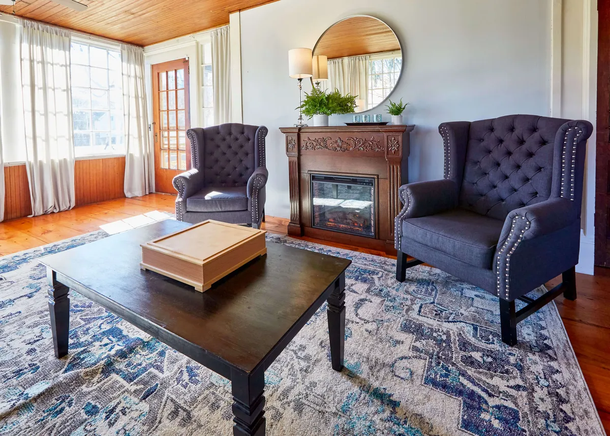 This image features two upholstered armchairs arranged by a fireplace, with a coffee table and decorative mirror completing the inviting setup. The room is filled with natural light, creating a cozy and welcoming ambiance ideal for socializing and gathering.