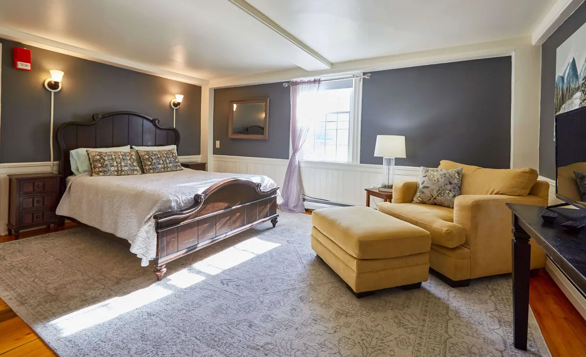 A spacious bedroom with a king-sized bed, an inviting armchair positioned near a large window, and vibrant artwork. The window draws attention to natural light and hints at scenic outdoor views, enhancing the connection to nature and the possibilities of local exploration.
