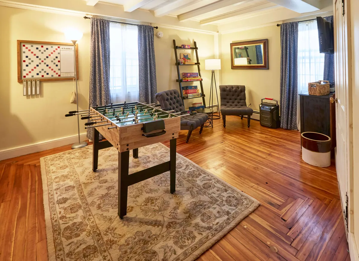 A cozy game room featuring a foosball table at its center, surrounded by comfortable chairs and a bookshelf. The room is warmly decorated with a soft rug, inviting players to enjoy both games and relaxation.