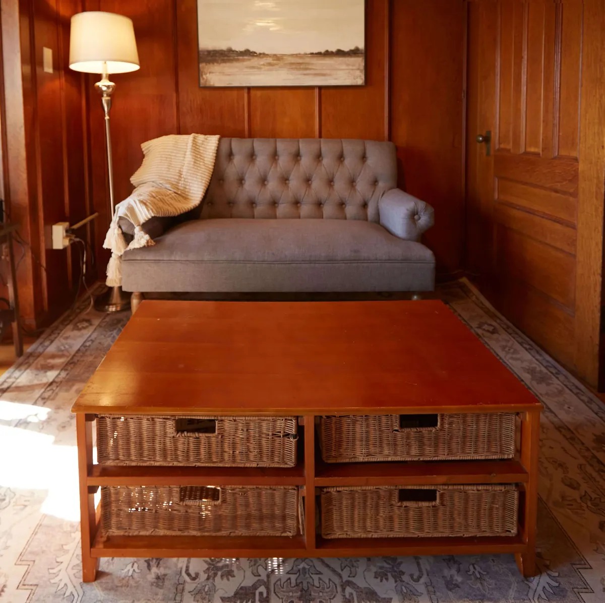 A gray tufted sofa sits beside a sturdy wooden coffee table with baskets, with a floor lamp providing soft illumination. The setup exudes comfort and is perfect for larger group conversations or intimate family moments.