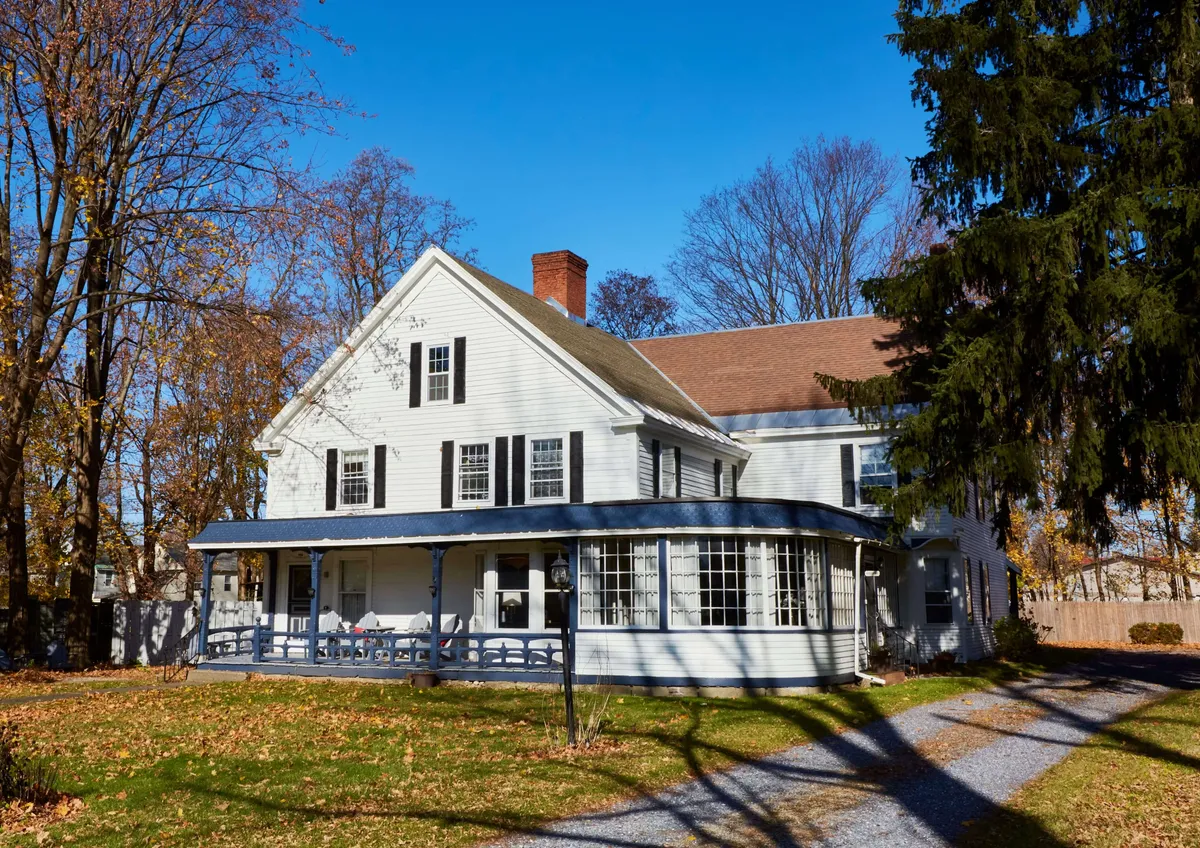 An inviting shot of the home's porch, large windows, and prominent chimney, framed by lush landscaping. The scene is serene, with hints of seasonal change and the promise of cozy gatherings both inside and out.