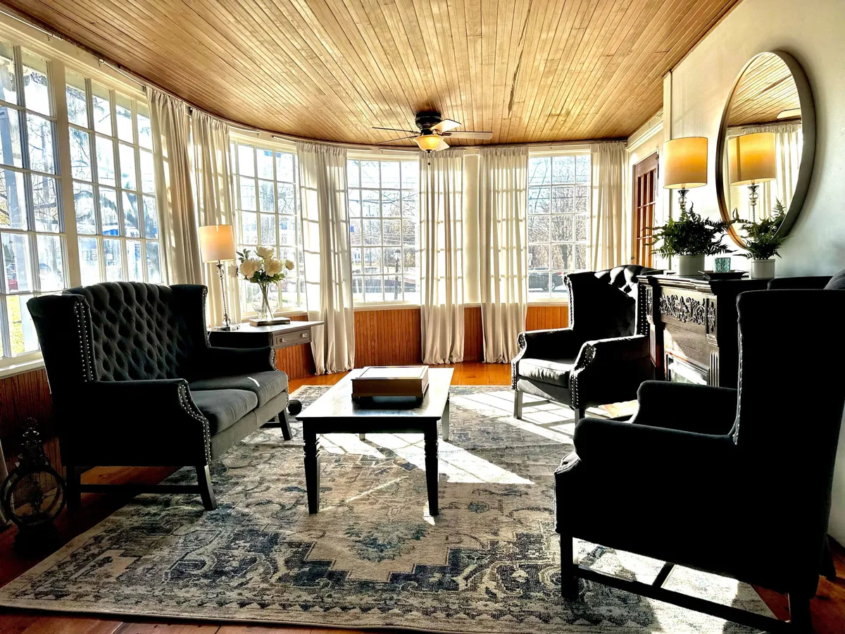 A bright and cozy living area with large windows, comfortable armchairs, a decorative coffee table, stylish area rug, and ambient lighting. The space is perfect for relaxed conversations, morning coffee, or hosting group gatherings in an elegant setting.